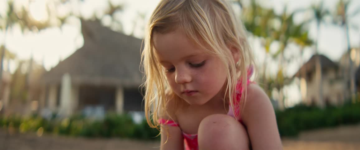 Blonde girl in a pink swimsuit looking down with buildings and palm trees in the background