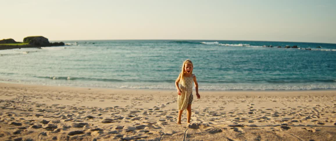 A young child walks barefoot across the sandy beach toward the ocean with gentle waves rolling in under a clear sky