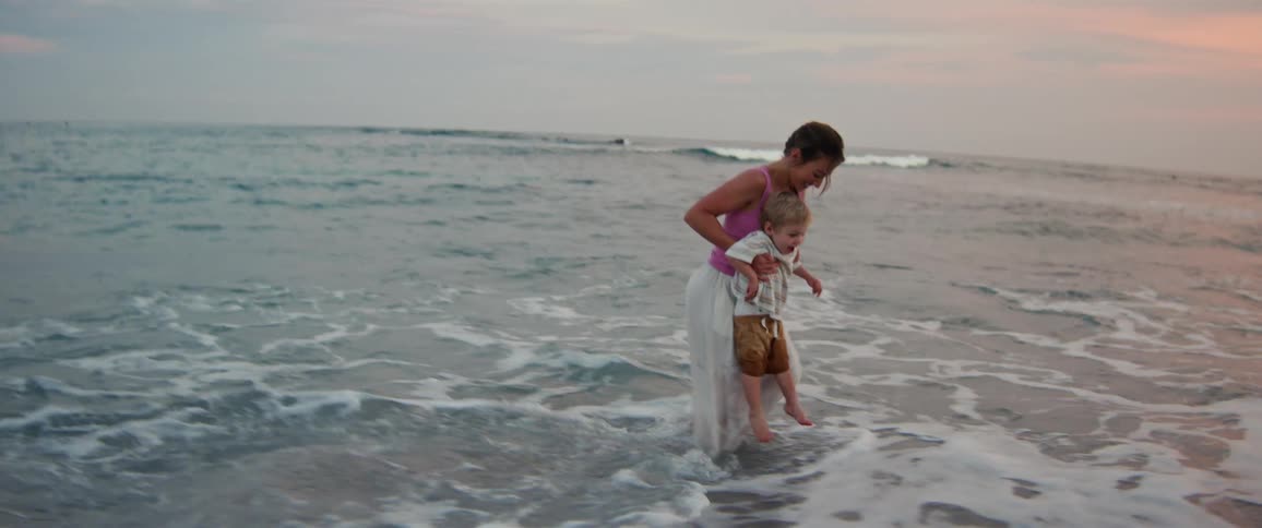 Little boy running barefoot on the beach into the water as waves roll in to shore as his mother picks him up and carries him away