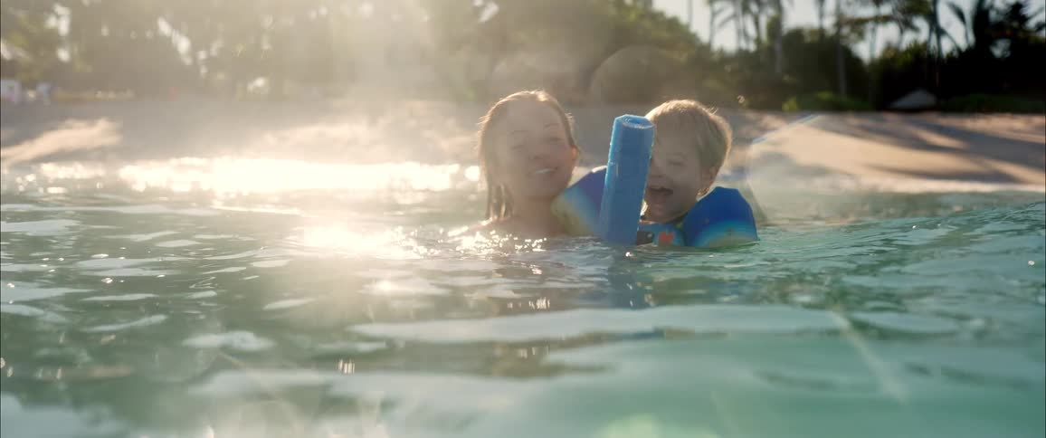 An adult and a young child laugh together while floating in the ocean sunlight sparkling on the water as they hold onto a blue pool noodle near the shore