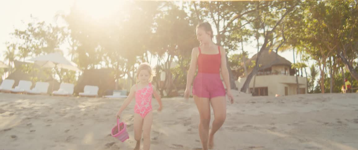 Woman and girl walking on the beach sand with a pink bucket at sunset