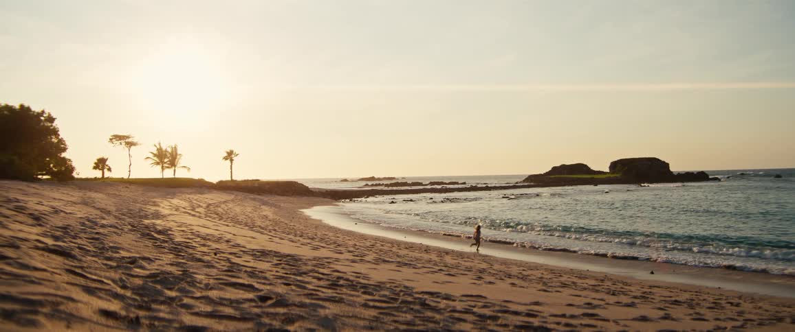 A young girl wearing a light dress runs across a sandy beach toward the ocean waves during a bright sunset with palm trees and rocky outcrops in the background