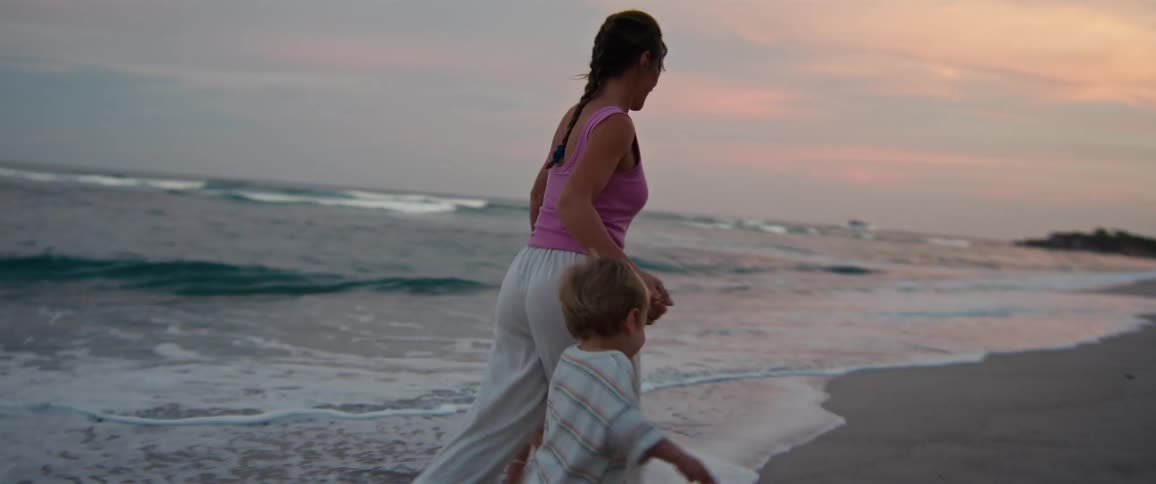 A mother and her two young children hold hands and run through the shallow water on a beach at dusk