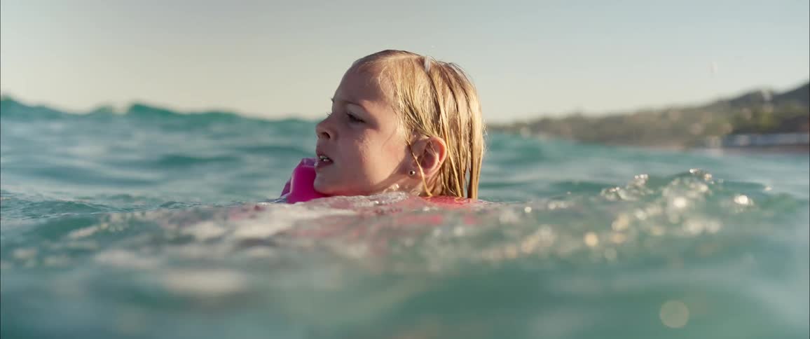 A young girl wearing a pink life vest swims through gentle ocean waves under a bright sunny skythe camera stays at water level capturing her expressive face as she navigates the swells near a distant coastline