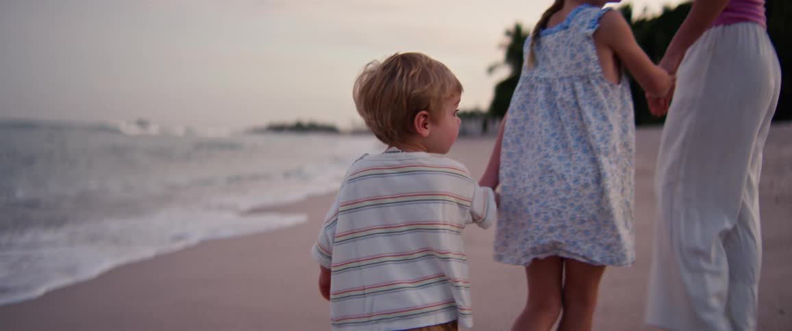 Boy walking on the beach sand while a girl holds his hand beside a woman