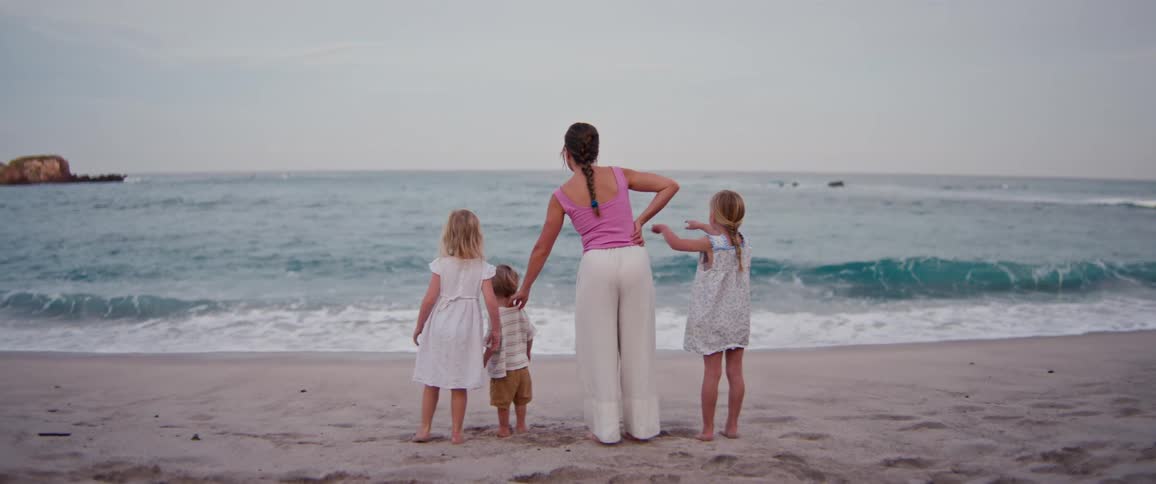 A woman and three young children stand on a sandy beach with their backs to the camera while watching the blue ocean waves under a cloudy sky