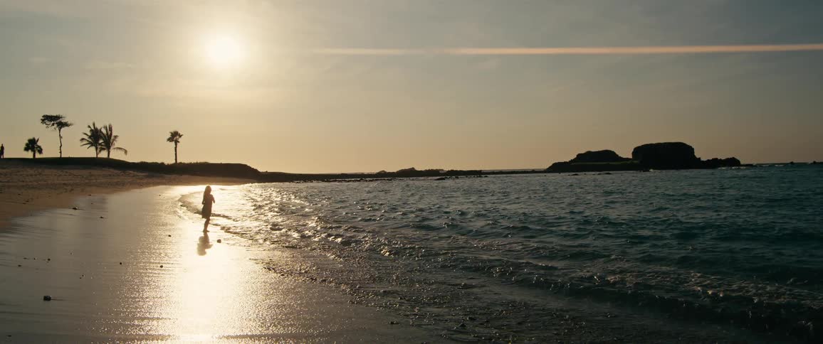 A young girl wearing a light dress stands in the shallow water of a sandy beach as gentle ocean waves roll onto the shore under a bright sun during a clear sunset