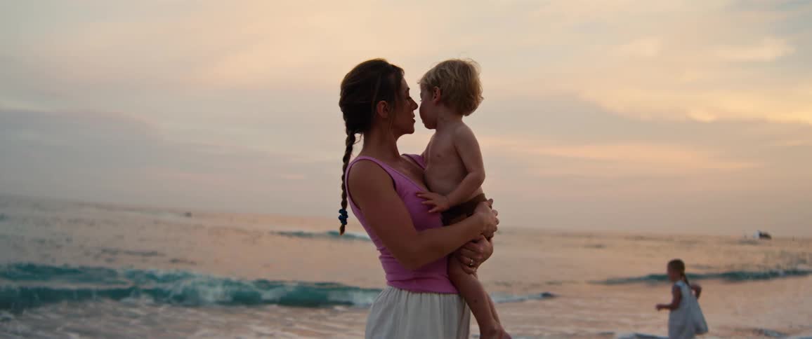 Woman holding a child in her arms on the beach at dusk while another girl walks on the sand in the background