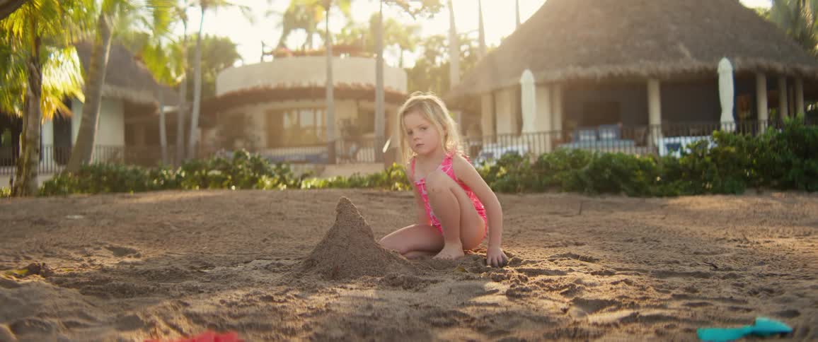 A young girl in a pink swimsuit sits on a beach at sunset carefully shaping a tall sandcastle with her hands while tropical buildings and palm trees stand in the soft backlight of the background
