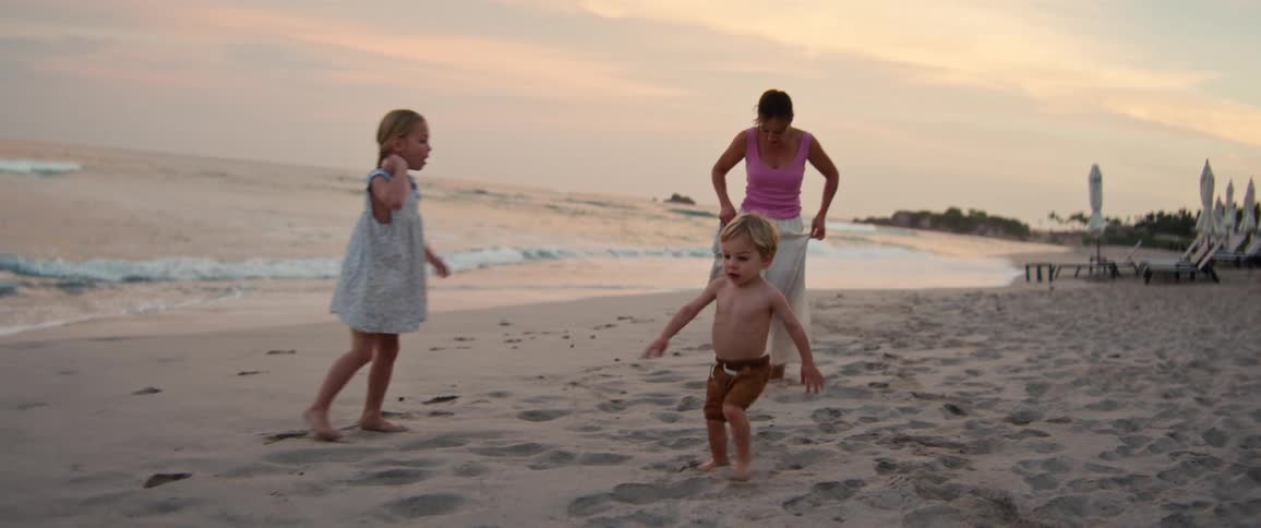 A young boy runs across a sandy beach at dusk while a woman and a girl watch him from the background near the crashing ocean waves under a hazy sunset sky