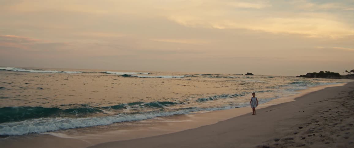 A young girl in a white dress walks slowly along the sandy shoreline as gentle ocean waves wash over the beach under a hazy sunset sky with distant rocks visible in the water
