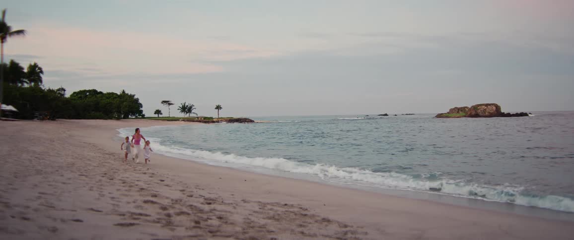Young adult woman running on the beach holding the hands of two young blonde girls outdoors during sunset time