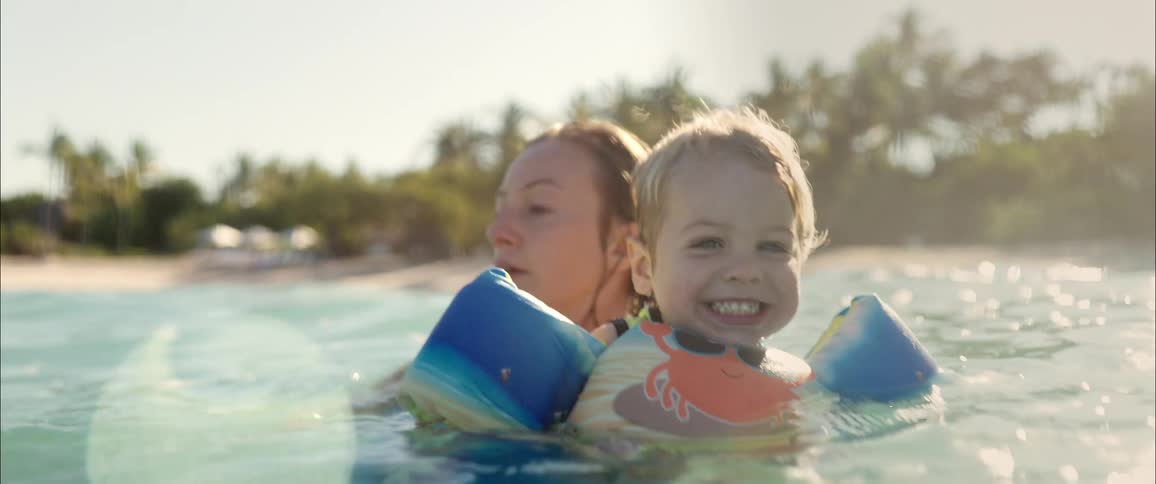 A woman supports a smiling young child wearing blue armbands and a crab life jacket as they float together in the shallow ocean water near a tropical beach with palm trees under bright sunlight