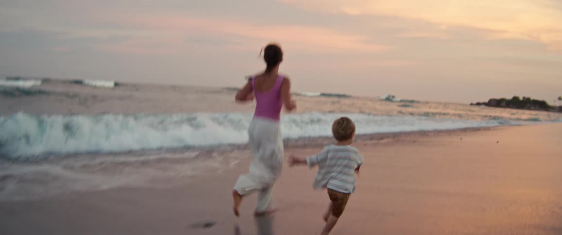 A woman in a pink top and white pants runs along the shoreline at sunset while holding hands with a small child before picking them up as they splash through the incoming ocean waves near a row of beach chairs