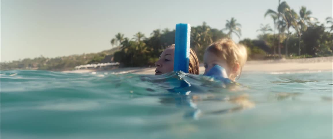 A mother and her young son enjoying a sunny day in the ocean near a tropical beach the mother supports the boy as he floats with the help of colorful arm dunkies and a blue pool noodle