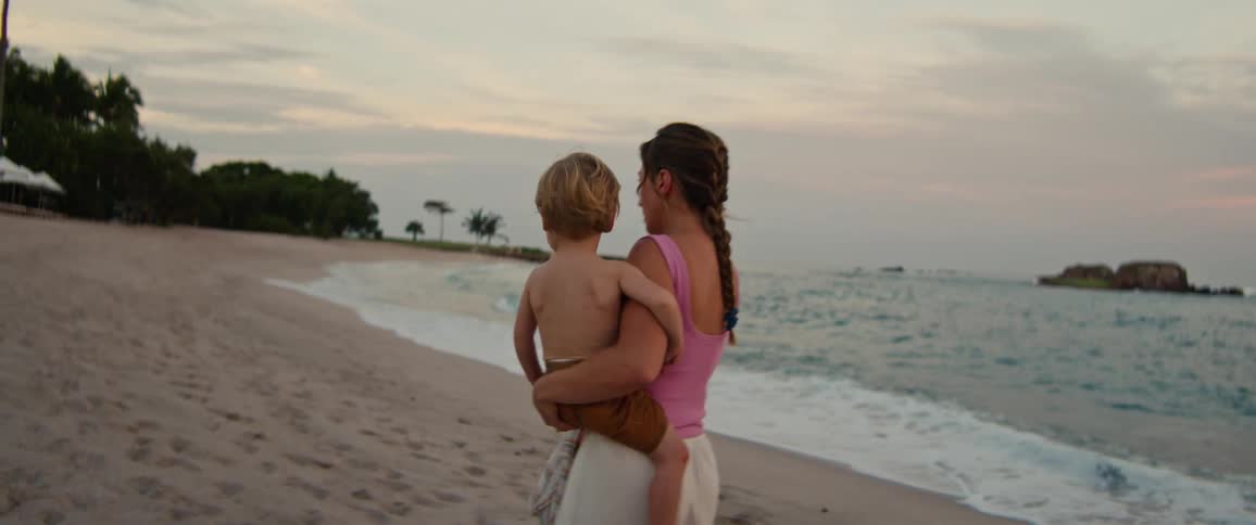 A woman wearing a pink tank top and white trousers carries a young child while walking along a sandy beach toward the ocean waves under a cloudy sky at dusk