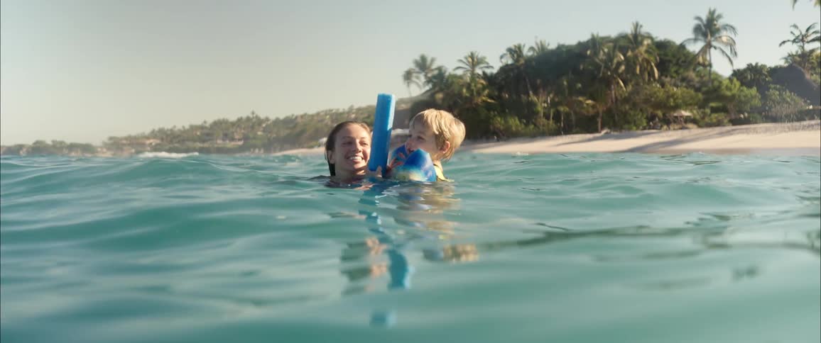 A smiling woman swims in turquoise ocean water while holding a young boy who wears blue arm floaties and holds a pool noodle with a tropical beach lined with palm trees and a sandy shore visible in the background under a bright sky