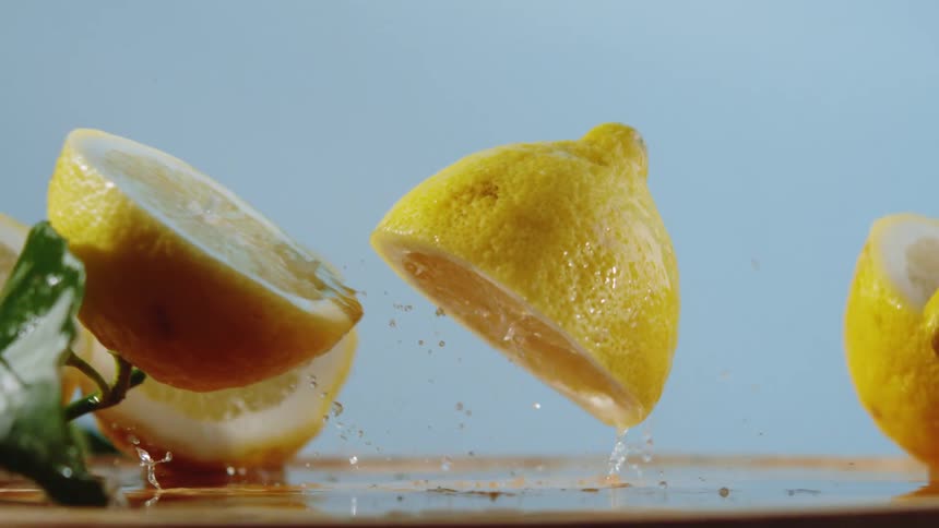 A bright yellow lemon with a green leaf attached is falling against a light blue background with sliced lemons arranged below