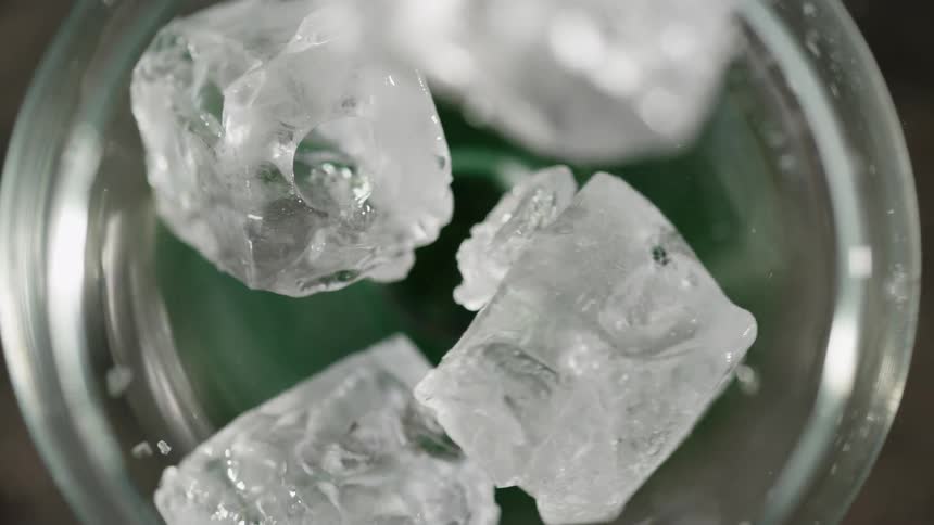 Ice cubes fall in slow motion into a glass filled with a green liquid the camera captures a topdown view as the ice splashes and settles at the bottom