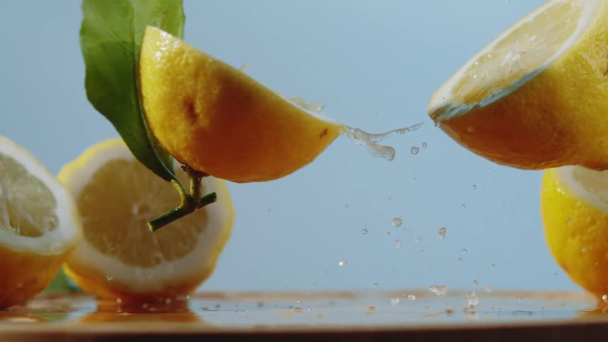A lemon falls from above in slow motion striking a flat surface and splitting open as it bounces slightly sending bright juice droplets spraying outward through the air before settling back down