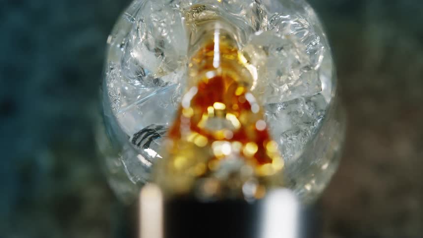 A closeup shot shows a golden liquid pouring from tip of a cup with a softly blurred circular light in the background