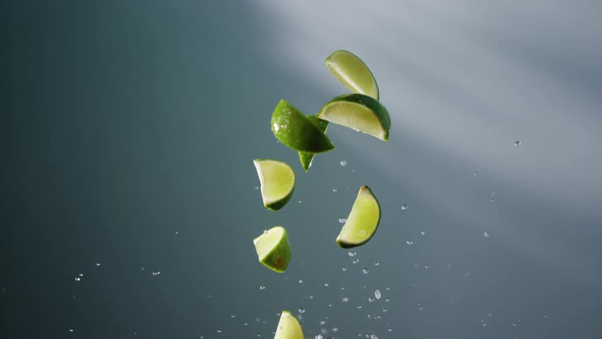 A slowmotion sequence of several lime wedges falling through the air against a muted bluegray background as the limes descend they are accompanied by a spray of water droplets that catch the light creating a refreshing and dynamic visual effect