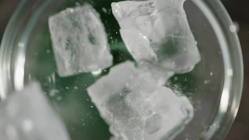 Ice cubes fall into the opening of a container with deep green liquid crashing around the rim