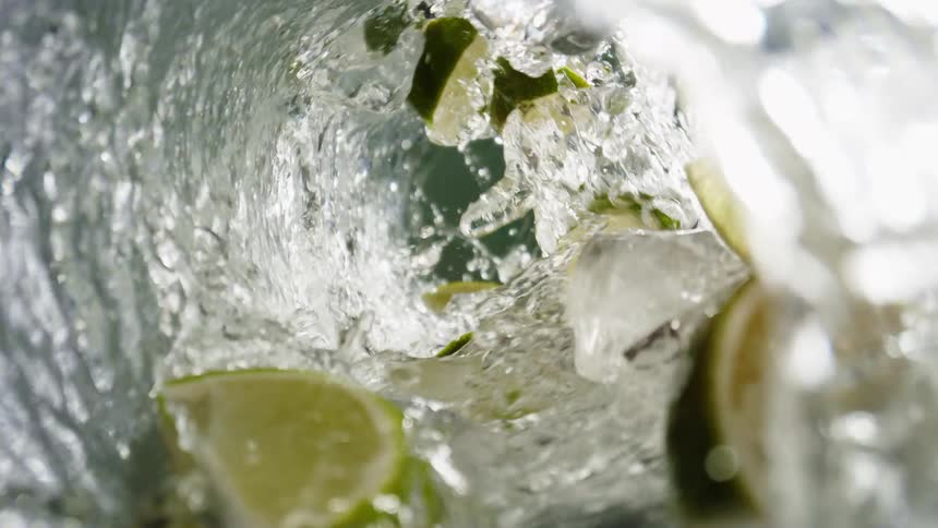 A view inside a rotating glass shows clear liquid ice cubes and lime slices swirling together to create a refreshing drink with a dynamic splashing effect