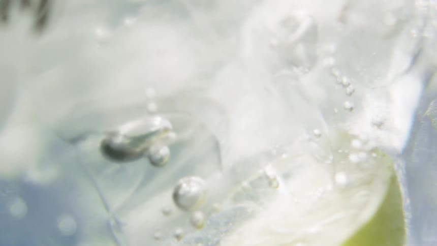 A lime slice falls into a glass of sparkling water and ice creating a splash and a stream of rising bubbles that swirl through the clear liquid in a crisp refreshing display