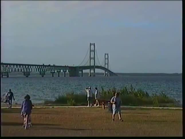 This video shows a wide view of a large suspension bridge spanning a body of water people are seen walking and relaxing in a grassy park area along the shoreline in the foreground
