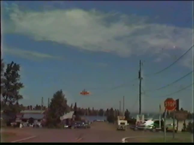 A view from a moving vehicle shows a road leading toward a body of water with trees and buildings under a blue sky with light clouds during a clear day