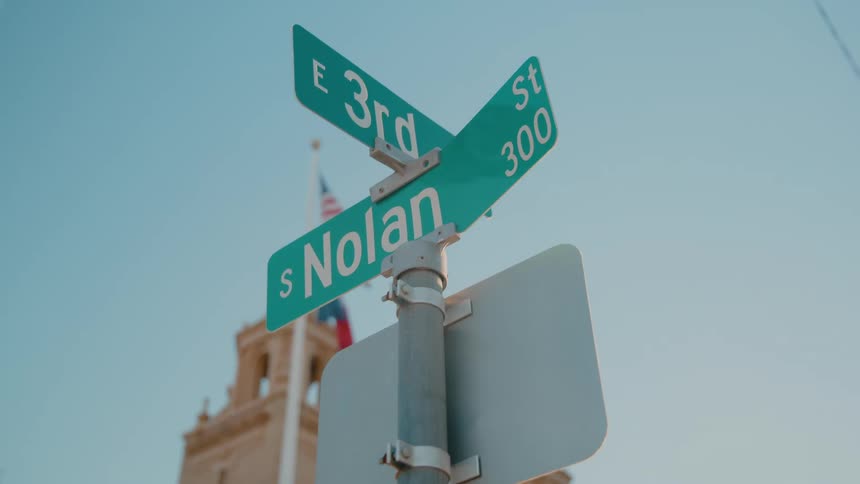 Green street signs mark the intersection of east 3rd street and south nolan street against a clear blue skya tall building with a flag and architectural detailing is visible in the blurred background behind the signpost