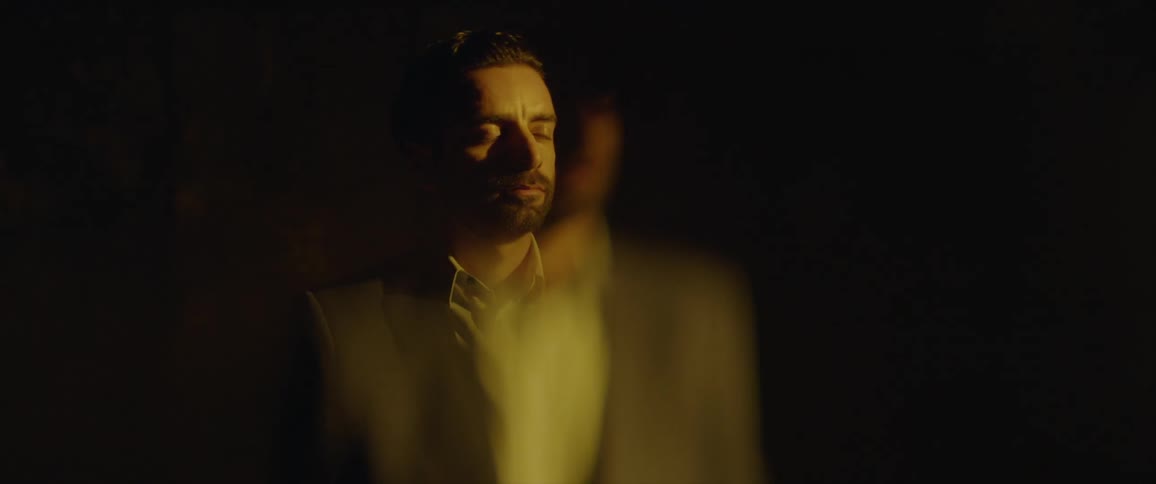 A man appears from the darkness and then closes his eyes as it goes dark again