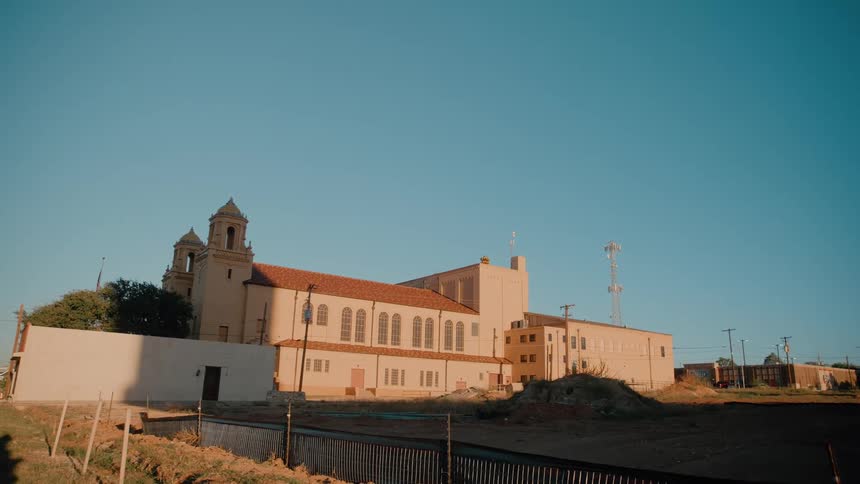 White church with towers and tall windows beside industrial buildings and a communication tower under blue sky