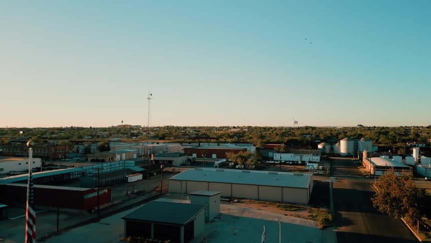This aerial footage shows a wide view of an industrial or smalltown landscape under a clear warm sky at sunset the camera pans slowly across the scene capturing various warehouses storage silos and a flagpole in the foreground