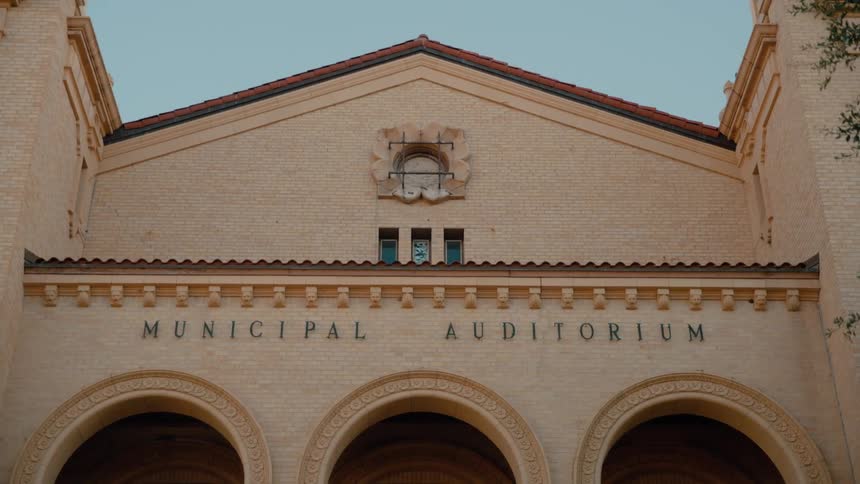 The front facade of a building features three large arches below the words municipal auditorium with a clear blue sky and a small tree branch visible at the edge of the frame