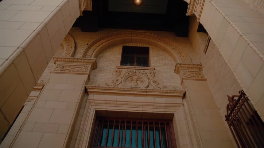 The camera tilts down the tan stone facade of a municipal building to reveal a set of large dark doors framed by ornate carvings and several decorative arches under a ceiling light