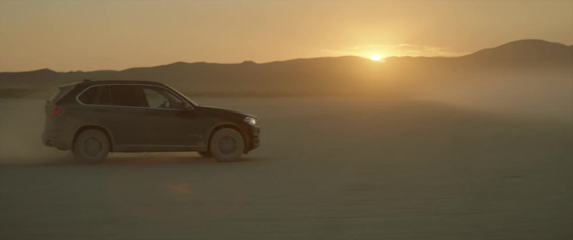 Vehicle moving across a sandy plain with the sun setting behind the mountains