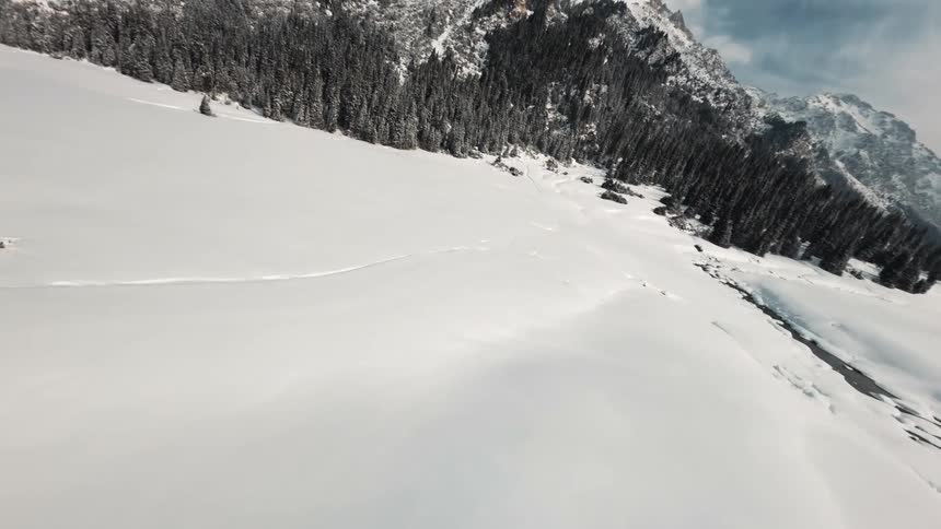 An aerial view shows a dark river winding through a wide snow covered valley with a dense evergreen forest along the banks and tall rugged mountains in the distance under a bright sky