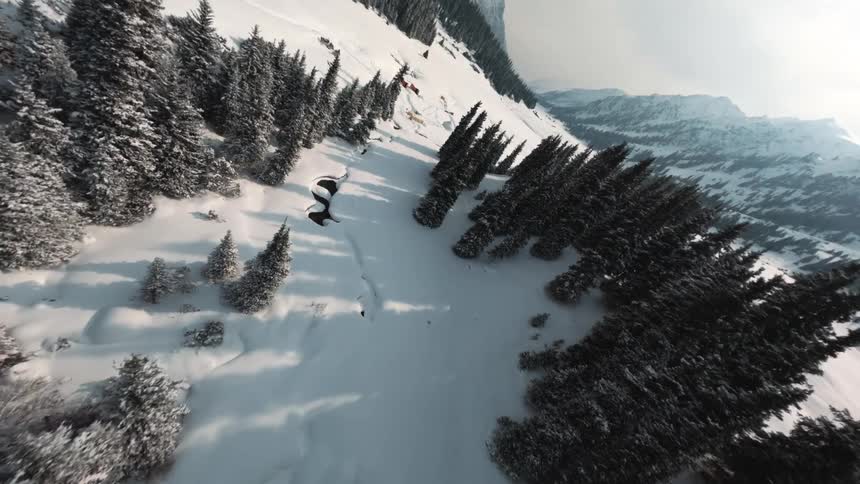 An aerial view shows a snowy mountain valley with dark green pine trees and small buildings under a bright sky