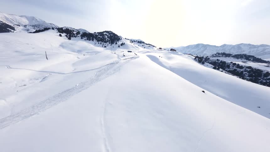 A view from above showing tracks on a snowy mountain slope with wooden fence posts and scattered trees under a bright blue sky