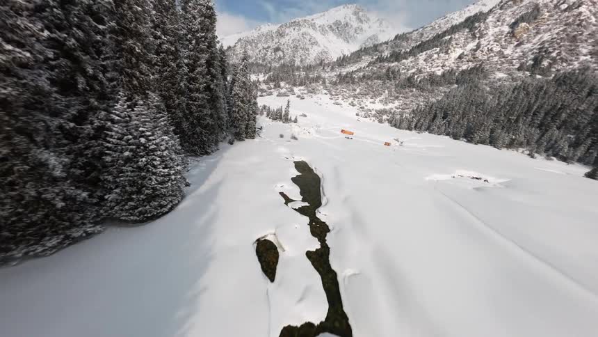 A high angle perspective of a dark winding stream cutting through deep snow between dense pine trees and distant mountain peaks under a blue sky in a remote winter valley