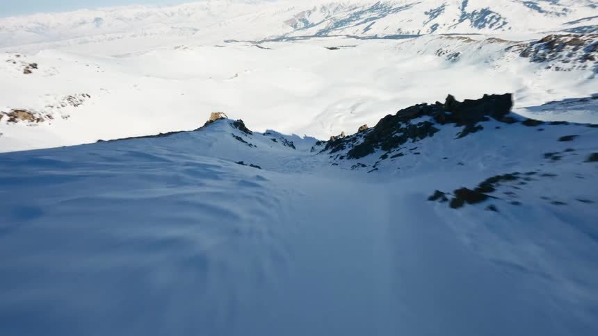 Acamera glides rapidly down a steep snowy mountain ridge toward a wide white valley surrounded by distant peaks under a bright clear sky