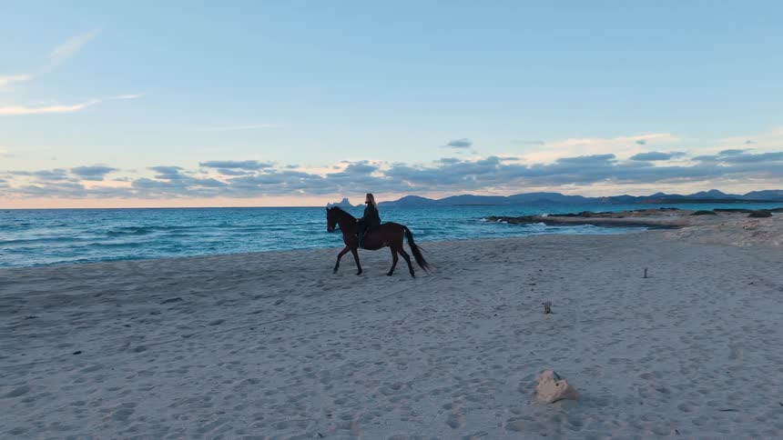 A woman wearing dark clothes rides a brown horse across a wide sandy beach next to blue ocean waves under a twilight sky with distant mountains