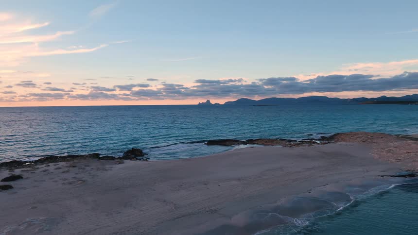 Beach with blue sea rocky formations and mountains on the horizon under cloudy sky