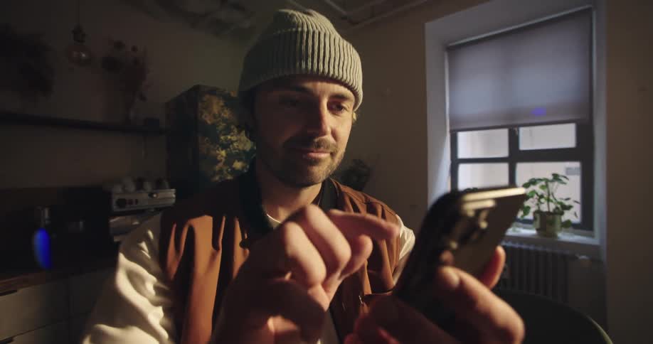 Fast dynamic push in of a man sitting on in a rustic room, tapping the screen of his smart phone and smiling