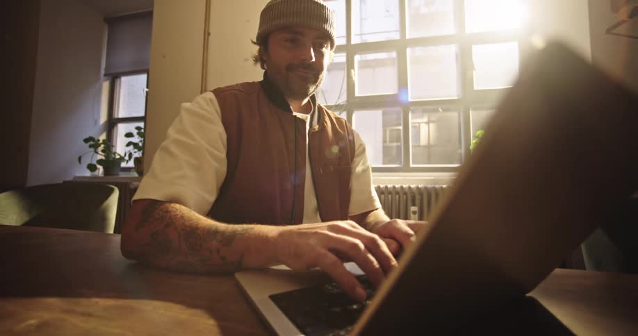 Man wearing beanie starting to work on his laptop at wooden table with sunlight coming through the window
