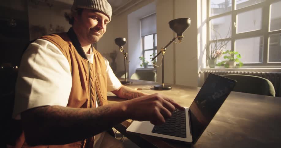 Dynamic shot of a man wearing a beanie and a tan vest sits at a wooden table in a sunlit room while opening his laptop and beginning to type