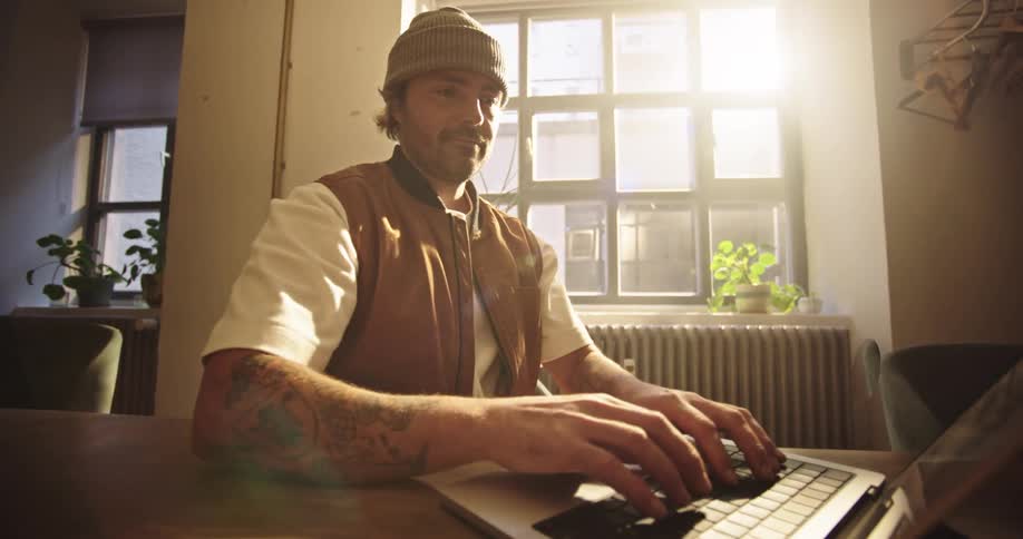 Dynamic shot of a man wearing a beanie and a tan vest sits at a wooden table in a sunlit room while opening his laptop and beginning to type