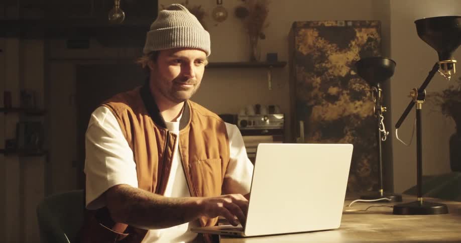 A man in an outdoor outfit types on a laptop while sitting at a table in a room